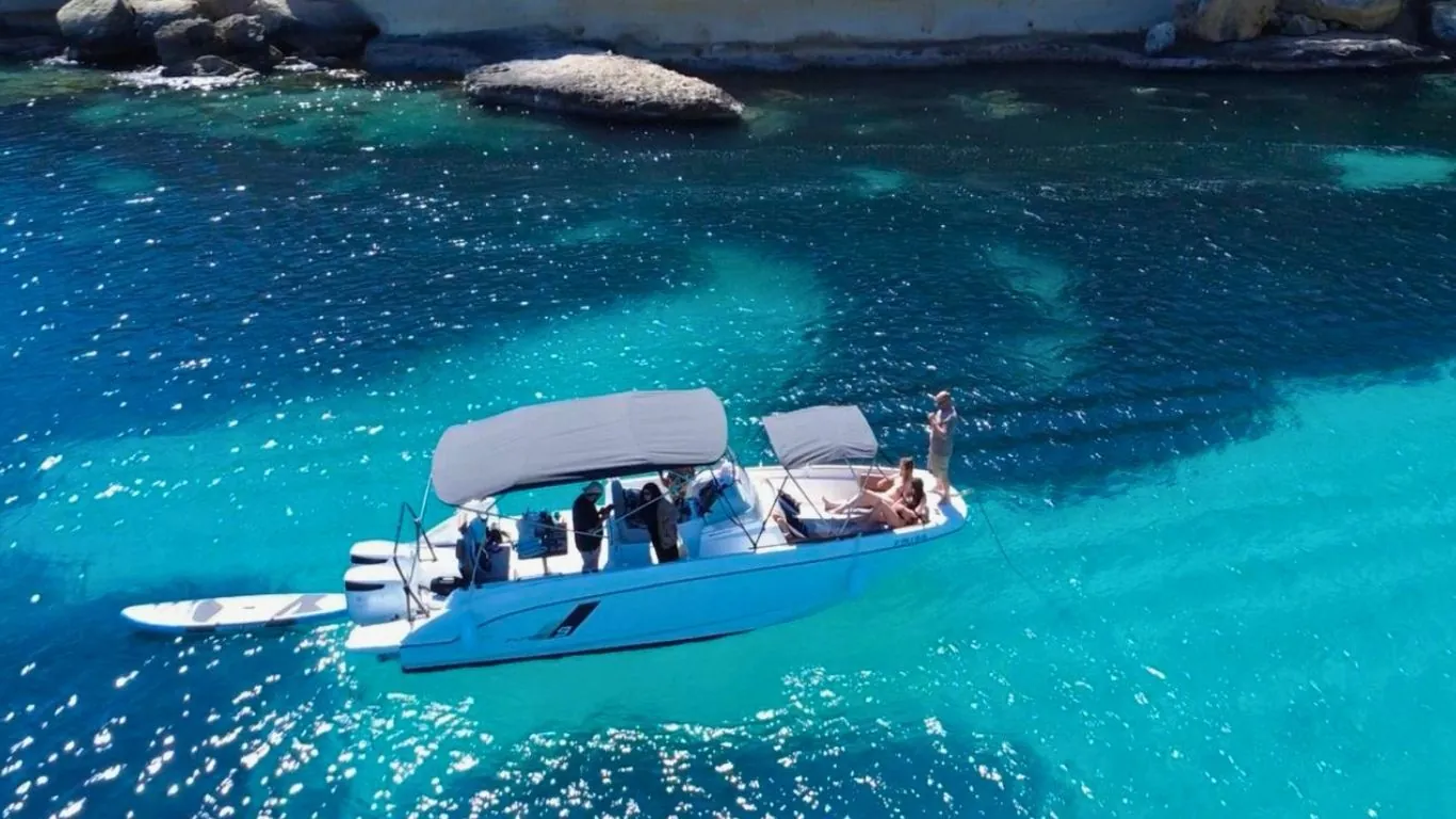 Barca de Holiboats fondeada en una cala de aguas turquesas en Mallorca, con invitados a bordo y una tabla de paddle surf en el agua